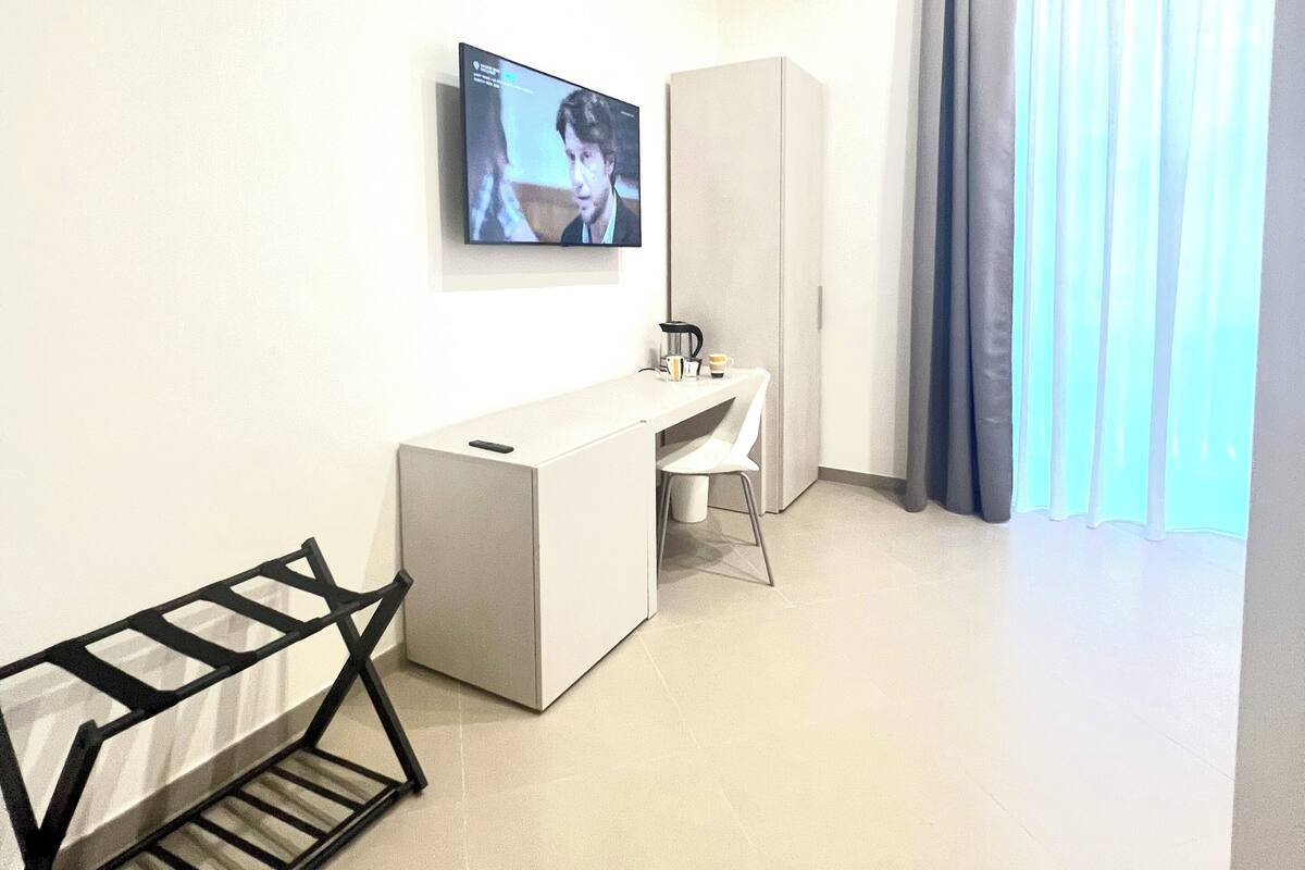 A modern hotel room with a flat-screen TV mounted on the wall, a white desk, a chair, a small cabinet, and a window with curtains.