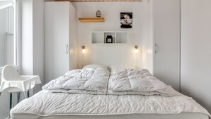 A bedroom with a bed, a chair, a shelf with decorative items, and a wall-mounted light fixture.