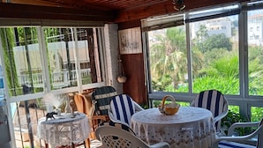 Um alpendre coberto com uma mesa redonda e cadeiras, um candeeiro pendente e grandes janelas com vista para o verde e edifícios.
