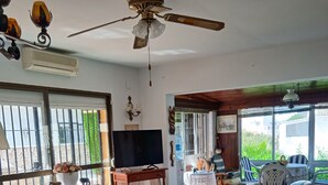 Sala de estar com TV, sofás de couro, ventilador de teto e vista através de amplas janelas.
