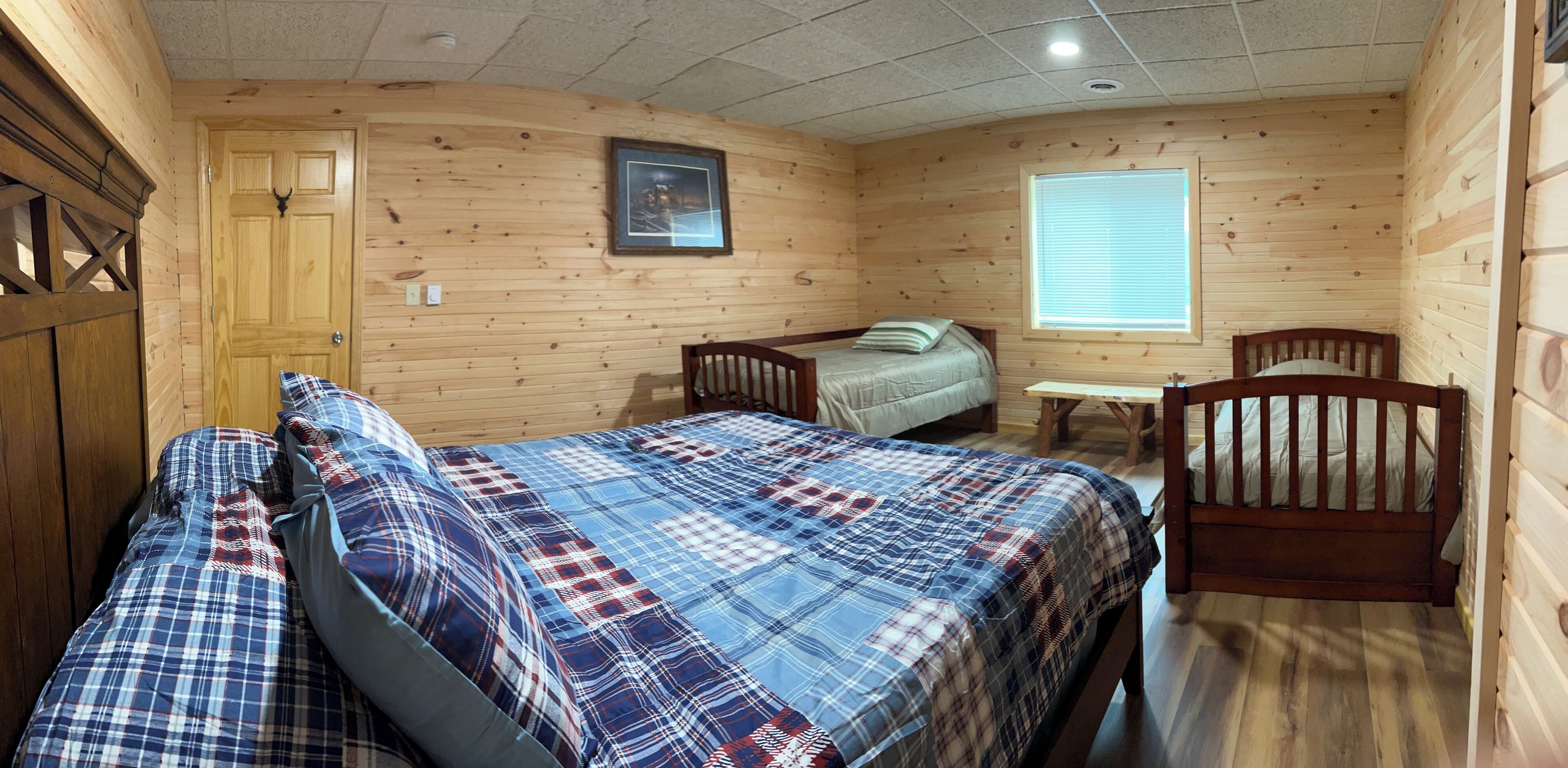 A room with wooden walls, a bed with a plaid bedspread, a small table, and a window with blinds.