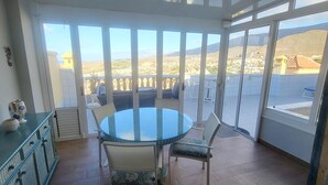 Uma área de refeições com mesa de vidro redonda e cadeiras, janelas amplas com vista para o horizonte urbano e colinas, e um armário verde-água com objetos decorativos.