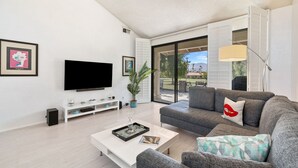 Un salon moderne doté d’un téléviseur à écran plat, d’un canapé gris, d’une table basse blanche et d’une grande fenêtre donnant sur l’extérieur.