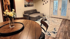 Un comedor con una mesa redonda de madera, un jarrón con flores, un sofá seccional gris, una alfombra estampada y un tronco de árbol grande de madera.
