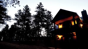 Un edificio de dos pisos con fachada oscura y ventanas iluminadas, rodeado de altos árboles.