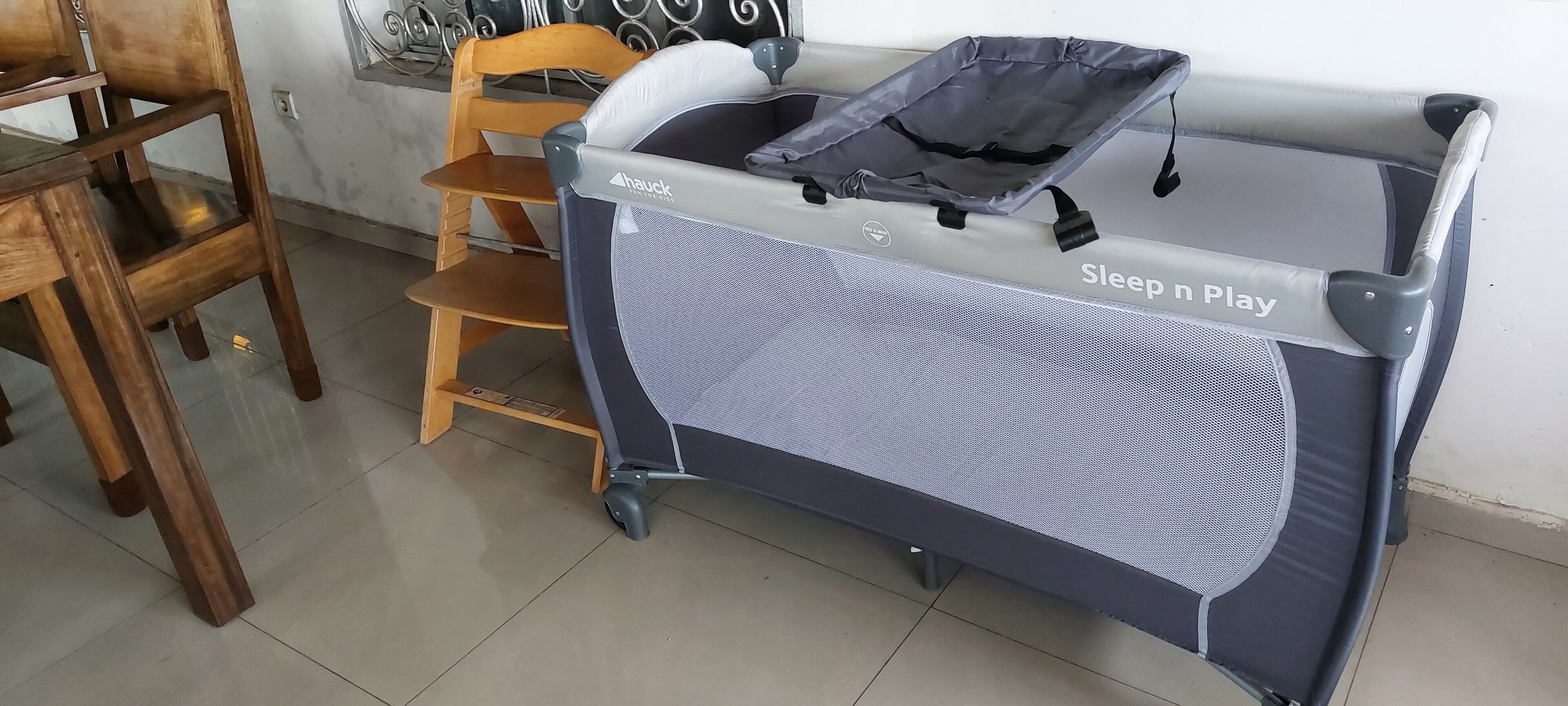 Un berceau portable portant la mention « Sleep n Play », une chaise en bois et un sol carrelé.