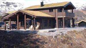 Puinen mökki, jossa on olkikatto, suuret ikkunat ja parveke, ja taustalla kohoaa vuoristo.