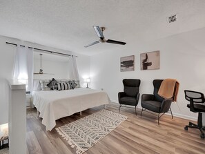 Une chambre à coucher comprenant un lit, des tables de chevet, un ventilateur de plafond, deux chaises, un tapis et des œuvres d’art accrochées au mur.