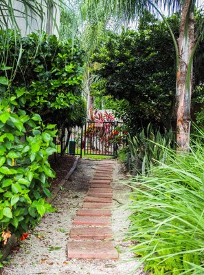 Un sentier de jardin qui mène à une zone délimitée par une clôture, où la végétation est luxuriante et où l’on trouve une grande variété de plantes.