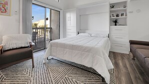 A bedroom with a bed, a chair, a sofa, a large window, and a balcony.