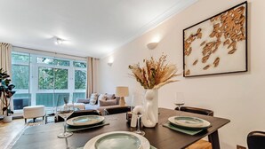 Een eetruimte met een donker houten tafel, gedekt voor vier personen, omringd door stoelen. Op de tafel staat een vaas met gedroogde planten, en erboven hangt een lijst met een kunstwerk aan de muur.