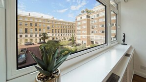 Uitzicht vanuit een raam op een meerlaags woongebouw met balkons, een kleine tuin en een standbeeld op de vensterbank.