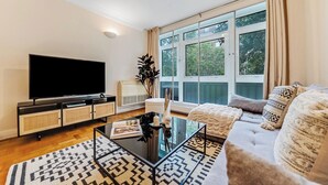 Een moderne woonkamer met een flatscreen televisie, een glazen salontafel, een bank en een groot raam met gordijnen.