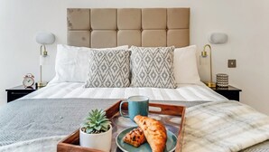 Een net opgemaakt bed met opvallende kussens, een nachtkastje met een lamp, een dienblad met croissants en een mok, en een kleine potplant.