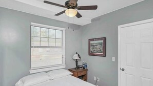 Un dormitorio con cama, ventilador de techo, ventana con persianas, mesita de noche con lámpara, un cuadro enmarco y una puerta.