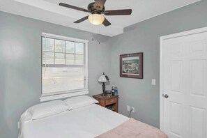 Un dormitorio con cama, ventilador de techo, ventana con persianas, mesita de noche con lámpara, un cuadro enmarco y una puerta.