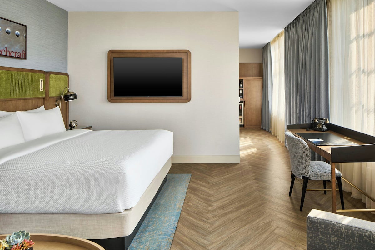 A hotel room with a large bed, a desk with a chair, a television, and a view of the city.