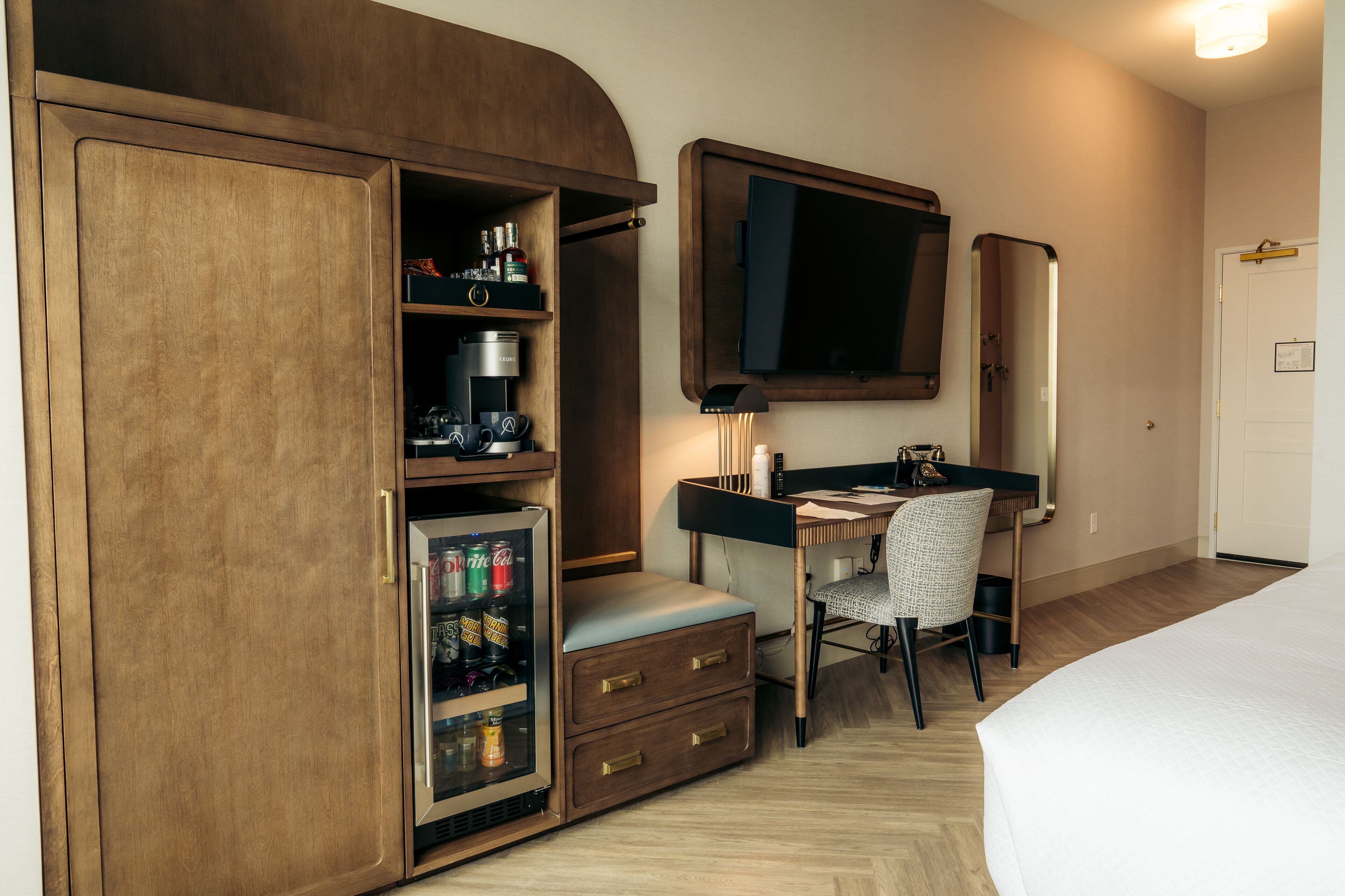 A compact hotel room with a bed, a desk with a chair, a wardrobe, a minibar, a television, and a mirror.