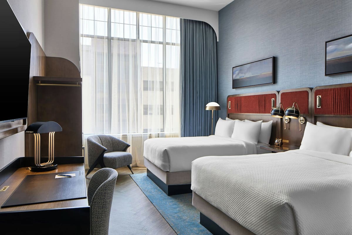 A hotel room with two beds, a desk, a chair, a lamp, and a large window with curtains.