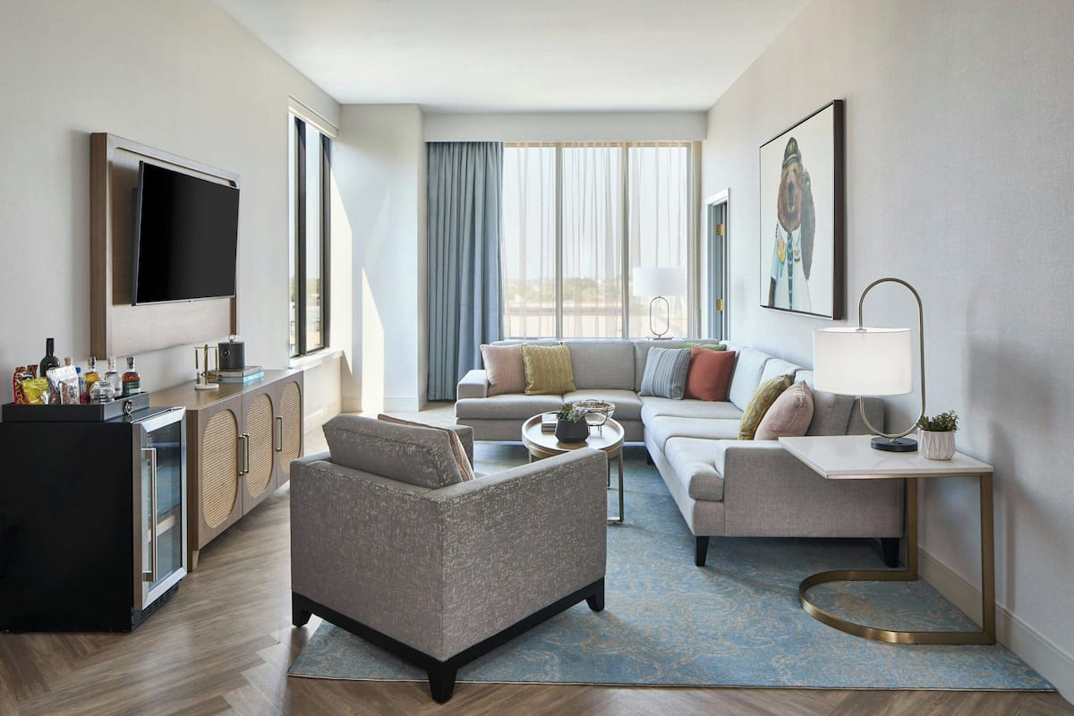 A modern hotel room with a flat-screen TV, a sofa, a coffee table, a side table with a lamp, and a minibar.