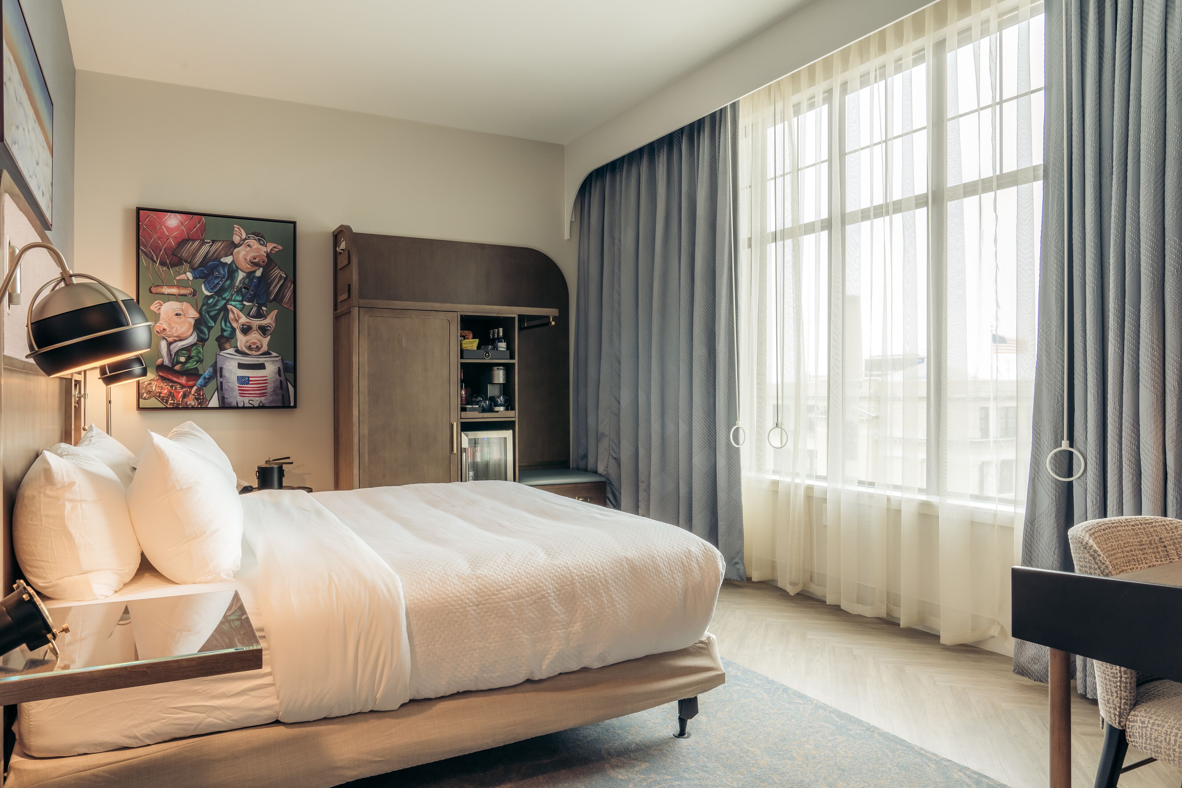 A hotel room with a large bed, a desk with a lamp, a painting on the wall, a wardrobe, and a large window with curtains.
