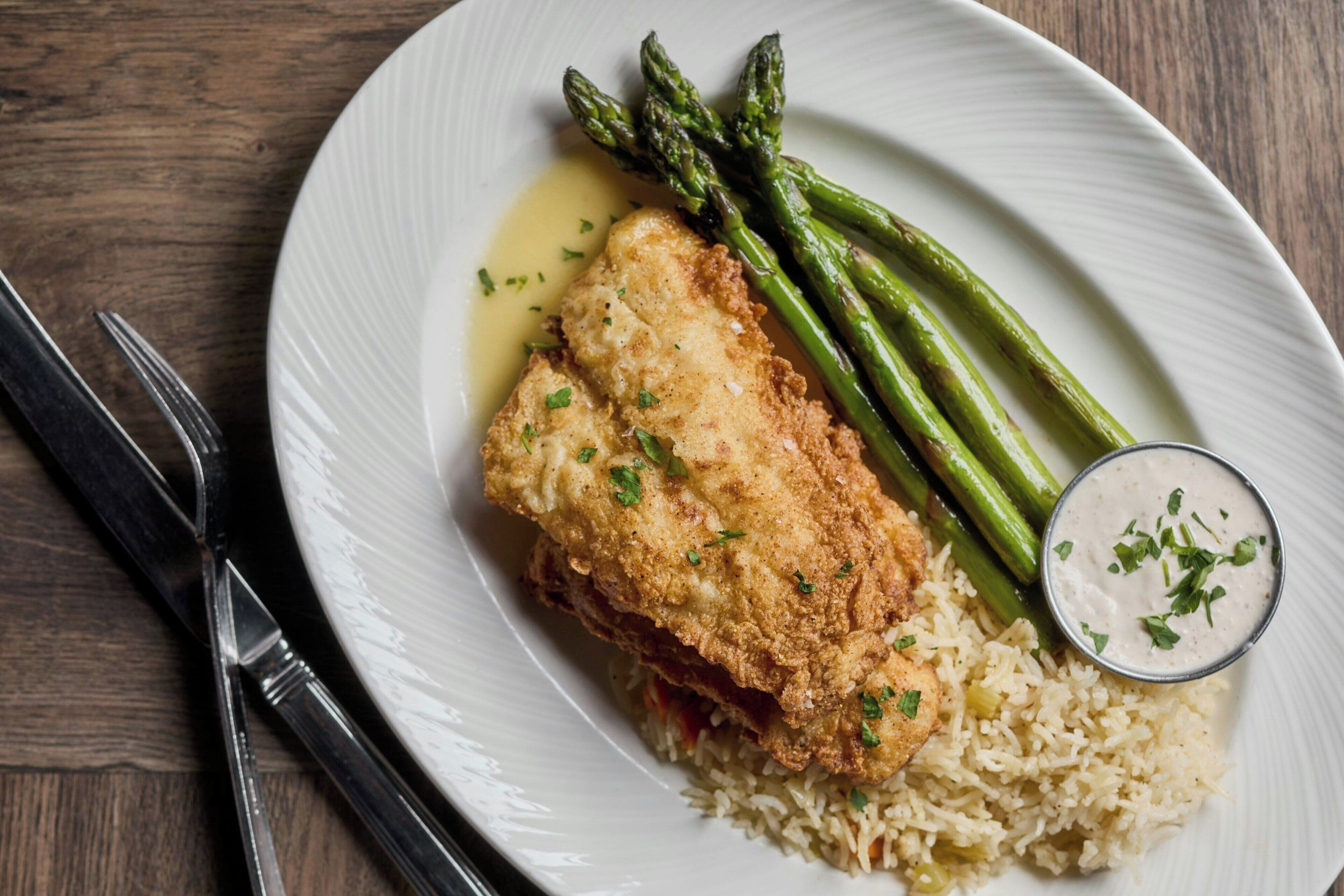 A plate with a breaded and fried item, asparagus, and rice, accompanied by a small bowl of sauce.