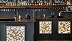 A bar area with a modern chandelier, decorative wall panels, and a variety of bottles and glasses on the counter.