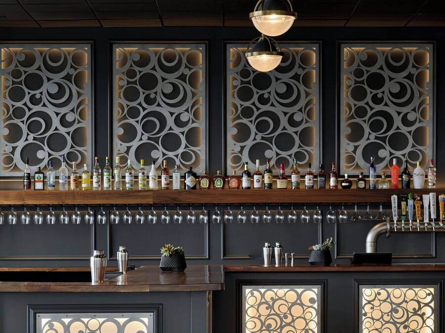 A bar area with a modern chandelier, decorative wall panels, and a variety of bottles and glasses on the counter.