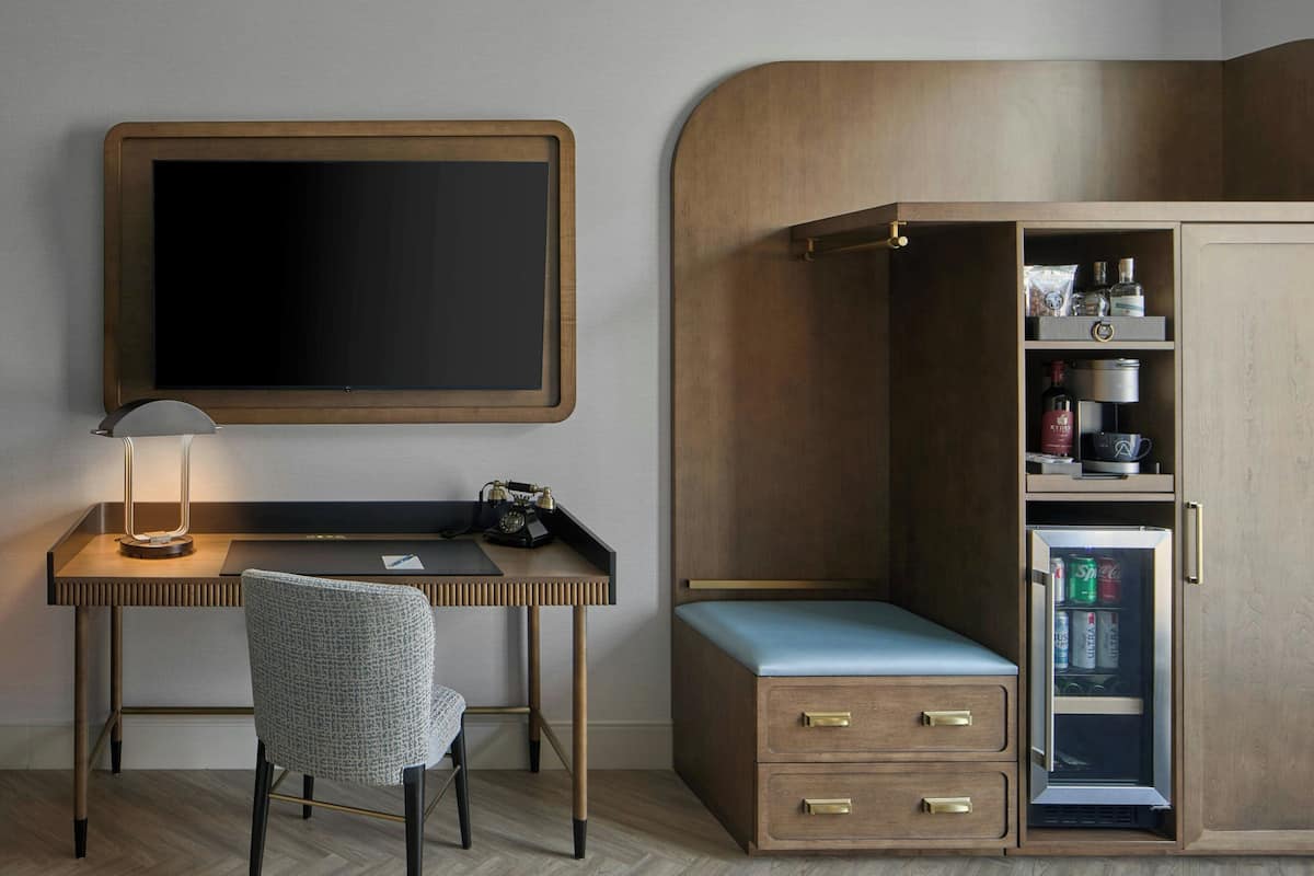 A compact hotel room with a wooden desk, a chair, a flat-screen TV, a minibar, and a wardrobe.