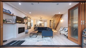 Una sala de estar moderna con chimenea, sofá y mesa de centro. Se observa una cocina al fondo y una escalera que sube a otro nivel.