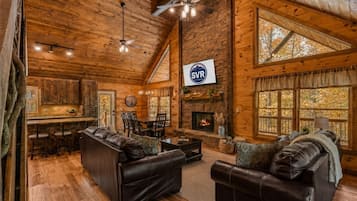A log cabin interior with a fireplace, leather sofas, a dining area, and large windows.