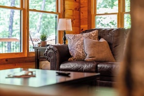 A cozy living room with a leather sofa, a side table with a lamp, and a view of the outdoors through large windows.
