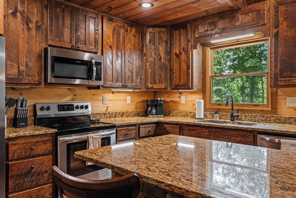 A kitchen with wooden cabinets, stainless steel appliances, granite countertops, and a window with a view of trees.