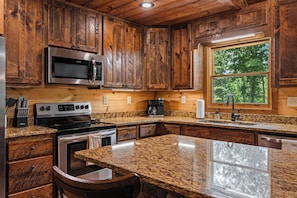 A kitchen with wooden cabinets, stainless steel appliances, granite countertops, and a window with a view of trees.