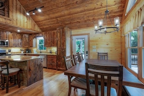 A rustic cabin interior with a wooden dining area, kitchen, and large windows.
