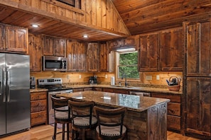 A kitchen with wooden cabinets, stainless steel appliances, a granite countertop, and a central island with bar stools. - Sunrays - Hot Tub, Outdoor Fireplace & Game (Morganton)