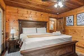 A wooden cabin-style bedroom with a large bed, a nightstand with a lamp, and a ceiling fan. - Sunrays - Hot Tub, Outdoor Fireplace & Game (Morganton)