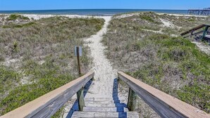 En träpromenad som leder till en strand med sanddyner och gles vegetation.