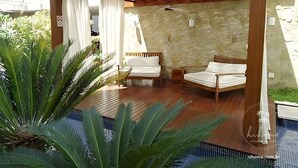 A covered outdoor seating area with two wooden chairs and a table, surrounded by lush greenery and a stone wall. - hihome – Mandai Flat (Cabo Frio)