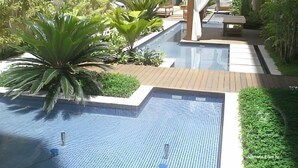 A modern pool area with a wooden deck, surrounded by lush greenery and a covered seating area. - hihome – Mandai Flat (Cabo Frio)