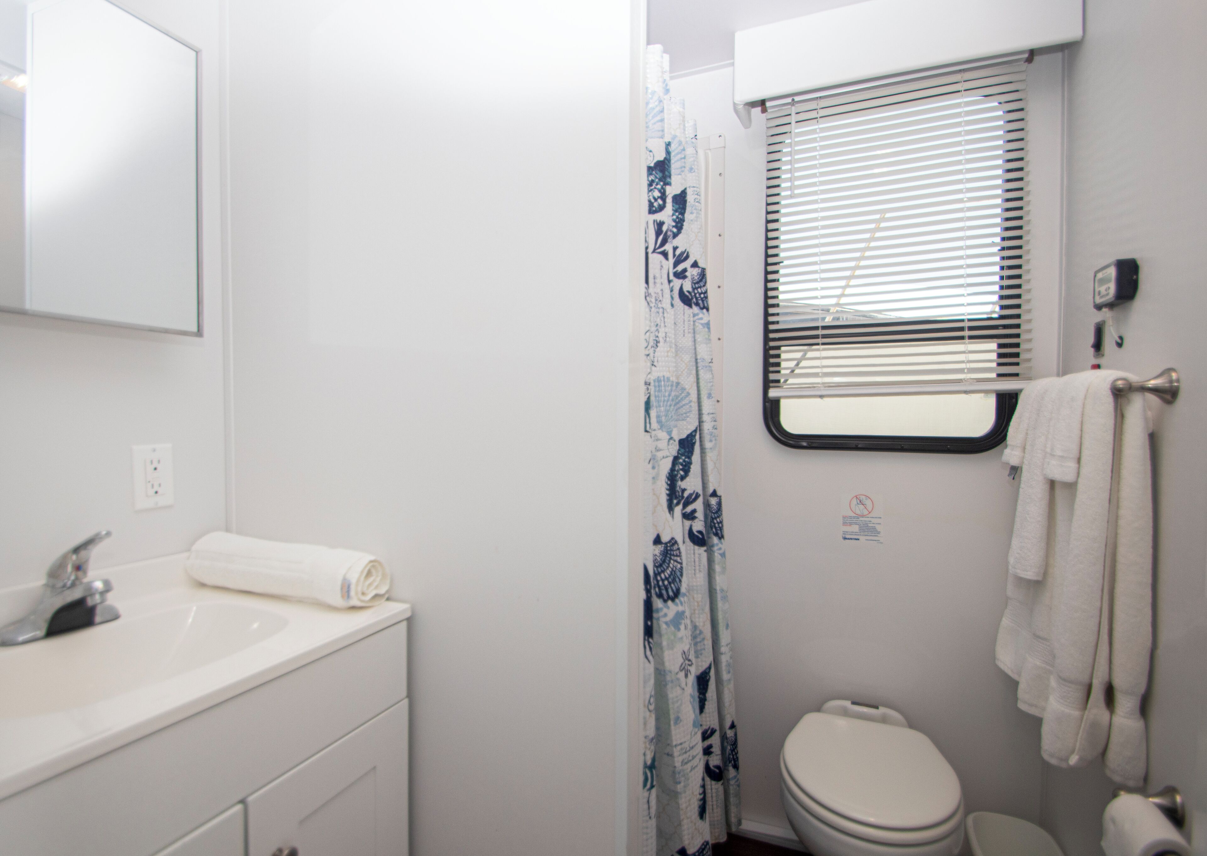 A bathroom with a white sink, a toilet, a shower curtain with a leaf pattern, and a window with blinds.