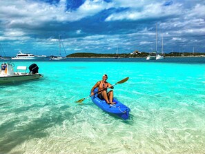 Une personne fait du kayak dans une eau turquoise et limpide, avec des bateaux et une île lointaine en arrière-plan.