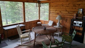 A cozy cabin interior with a round table, wicker chairs, a small lamp, and a bookshelf with books and a radio.