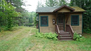 A small wooden cabin with a porch and steps leading to the door, surrounded by dense forest and a pond.