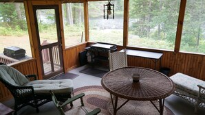 A wooden deck with a grill, a wicker chair, a table, and a view of the forest.