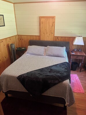 Una cama con cubrecama estampado y dos almohadas, una mesita de noche con lámpara, y un armario de madera en una habitación con paneles de madera.