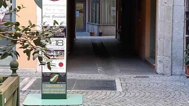 An outdoor area with a paved walkway leading to a building entrance. There is a signpost with text and a map, and a potted plant to the left.