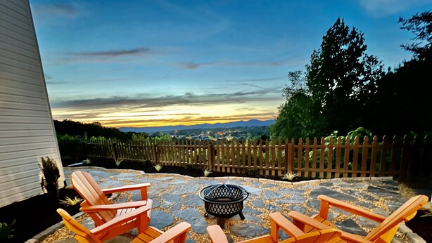 A patio with four Adirondack chairs, a fire pit, and a view of a sunset over a distant cityscape. - Iconic Dahlonega View! In Town! WINE GOLD HIKING (Dahlonega)