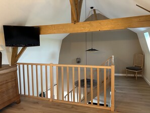 A loft-style room with wooden beams, a TV mounted on the wall, a staircase, and a chair. - The Pigeonnier, gîte at Domaine D'antan (Ennevelin)