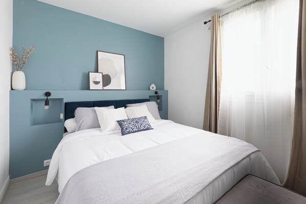 A modern bedroom with a bed, white bedding, a blue accent pillow, a framed artwork, and a vase with dried flowers on the wall. - Appartement Beauvais - Cocooning (Beauvais)